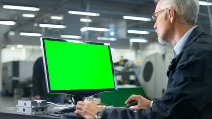 Senior engineer in glasses is working on a desktop computer with a green screen on monitor in a...