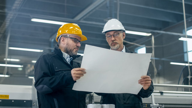 Two engineers in hardhats discuss a blueprint while standing in a factory.