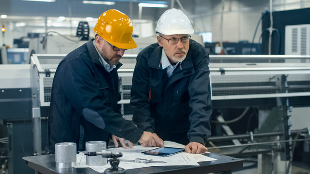 Two engineers discuss a blueprint while checking information on a tablet computer in a factory.