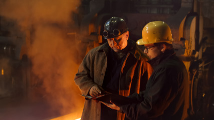 Fototapeta na wymiar Two Workers Have Conversation and Using Tablet PC in Foundry. Rough Industrial Environment.