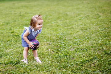 Little girl walking on the street. The kid is walking on the green grass. Girl with purple ball