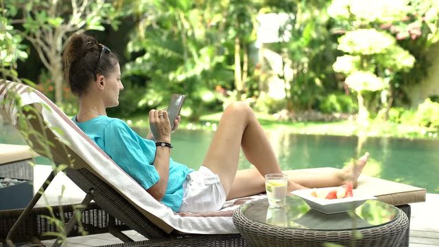 Woman relaxing on sunbed next to the swimming pool and browsing internet on smartphone, steadycam shot
