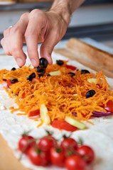 Preparing vegetarian burrito with vegetables and olives close up on wooden brown background. Hand with black olive. On blurred foreground small cherry tomatoes. Horizontal image. Side view