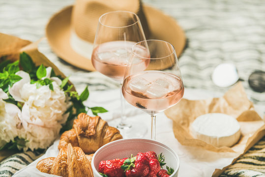 French style romantic summer picnic setting. Flat-lay of glasses of rose wine with ice, fresh strawberries, croissants, brie cheese, straw hat, sunglasses, peony flowers. Outdoor gathering concept