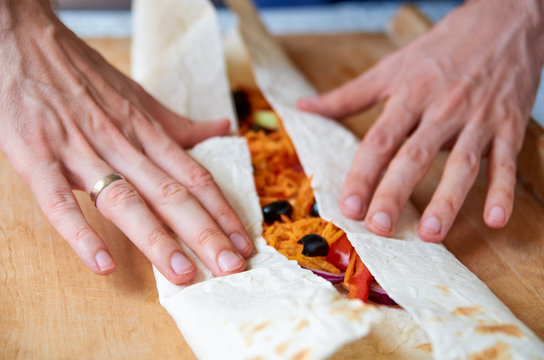 Man hands preparing food - vegetarian burrito with vegetables, olives, carrot, bell pepper and tomatoes close up on wooden brown background. Side view