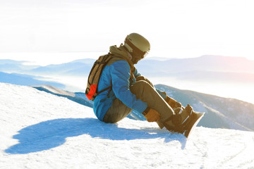 Young snowboarder in helmet sitting at the top of a mountain and tightening his bindings