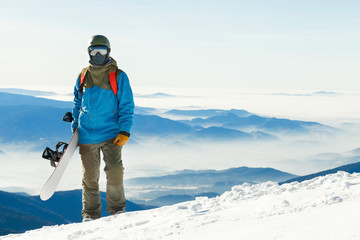 Fototapeta na wymiar Young snowboarder in helmet standing at the top of a mountain with beautiful sky on background
