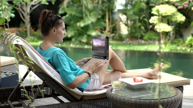 Woman using modern notebook while sitting next to the swimming pool, steadycam shot
