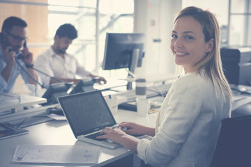 Business woman in office. Woman using laptop and looking at camera.