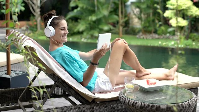 Woman watching something funny on tablet while relaxing next to the swimming pool, steadycam shot
