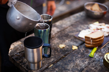 Man pour boiled water to hot mugs on wooden table during breakfast at the forest camp. People on picnic camping at national park and doing lunch tea and coffee. Isolated view