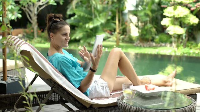 Attractive woman relaxing on sunbed and browsing internet on tablet, steadycam shot
