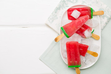 Frozen cold icy watermelon popsicles on wooden stick with green mint, basil leaves on ice cubes in oval plate, white background. Summer dessert ice cream, top view, flat lay, copy space for text