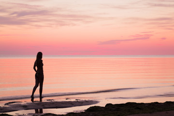 Fototapeta na wymiar Young woman walking alone at the seaside in summer evening. Woman silhouette on the beach. Sea water reflecting a pink heavenly colors and creating a romantic atmosphere.