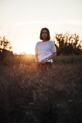 Naklejka na ściany i meble Young attractive brunette standing alone in the summer sunset field, enjoying her solitude and smiling