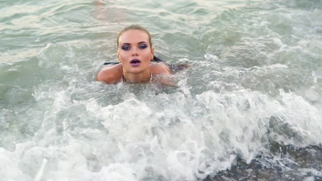 Sexy model in black leather bikini lying on the shore of the beach in the waves looking in the camera and flirting with the viewer. Attractive young woman with art golden makeup on the sand by the sea