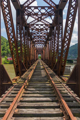 A down the track perspective across an old rusted railroad bridge spanning a river with green mountains in the distance with a blue cloudy sky.