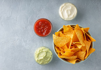 Bowl of crusty nachos or tortilla with different dipping sauces on gray stone background.