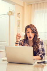 Shocked woman holding coffee cup and looking at laptop.