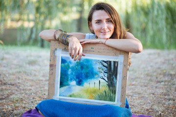 Portrait of charming female artist with picture and brush at the park