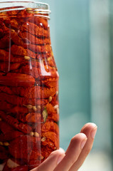Jar with many dried red tomatoes, olive and spices in man's hand with copy space on blurred nature green background close up. Dried tomatoes texture blurred background close up. Side view