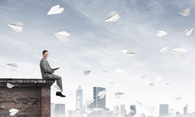 Man on roof edge reading book and cityscape at background