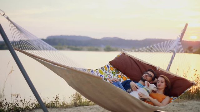 Young couple lying on a hammock with a dog. Feeling happy, smiling. Stroke the dog Near the lake