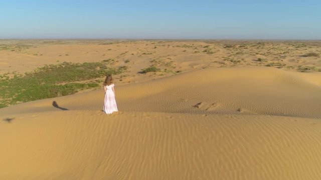 Young lonely woman with long hairs in white dress is staying on sand beige dune in desert. Aerial view.