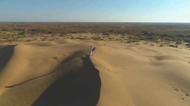 Young lonely woman with long hairs in white dress staying on the top of sand beige dune in desert. Drone is flying around. Aerial view.