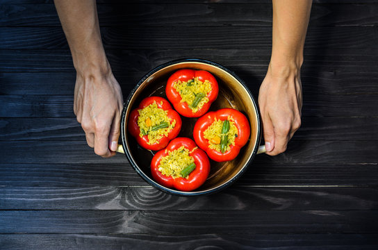 Chef holding a pan with four red stuffed peppers on dark rustic kitchen table background, top view. Cooking process