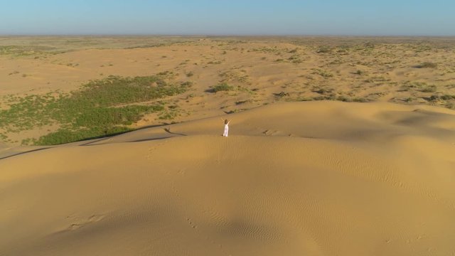 Young lonely woman with long hairs in white dress is staying with raised hands on sand beige dune in desert. Aerial view. Camera is tilting down.