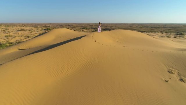 Young lonely woman with long hairs in white dress staying on the top of sand beige dune in desert. Drone is approaching. Aerial view.