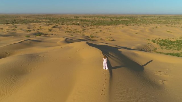 Young lonely, woman with long hairs in white dress is walking on sand beige dune in desert. Aerial view.