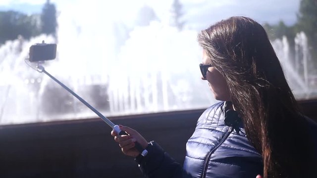 The girl makes a photo on the selfie stick, near the fountain, the hair develops downwind. slow motion. 1920x1080. full hd