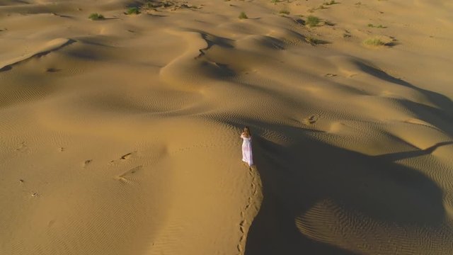Young lonely woman with long hairs in white dress is walking on a top of sandy beige dune in desert. Aerial view. Camera is tilting down..