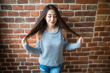 Beautiful young woman in casual wear hands play with hair smiling, standing against white brick wall