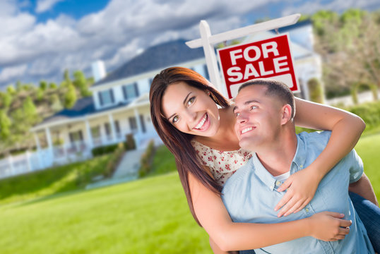 Playful Excited Military Couple In Front of Home with For Sale Real Estate Sign.