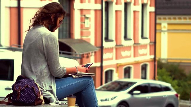 Video with young tourist woman sitting on parapet in city centre. Girl in casual clothes relaxing after long walk. Toned footage.