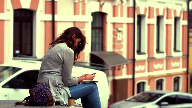 Tourist girl with curly hair taking rest after long walk. Traveling in the city, woman sitting on parapet in the street. Toned footage.