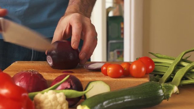 A man chopping, preparing a red onion on a wooden chopping board with a mixture of vegetables on the table. Dolly panning shot.