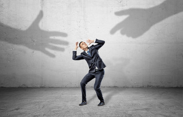 A scared businessman hides from two giant shadows of human hands reaching to him.