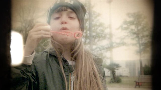 Fake 8mm amateur film: a little girl in a playground blowing into a plastic circle, trying to make soap bubbles. Outdoors shot, cloudy cold day. Glimmering strong light.
