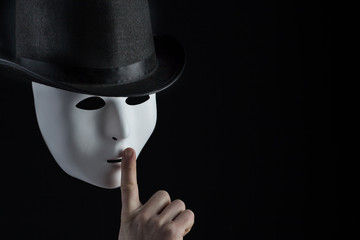 Male finger showing shh sign on white mask wearing black top hat on black background with copy...