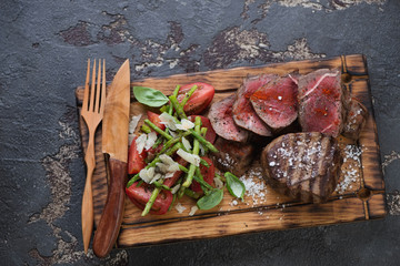 Grilled medallion beefsteaks with asparagus and tomato salad on a wooden serving tray. Flat-lay on a brown stone background, horizontal shot