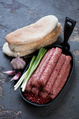 Frying pan with raw balkan cevapi or cevapcici sausages and pita bread on a brown stone background, studio shot