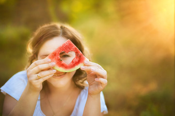 A girl in a white T-shirt is holding a dolm of watermelon with a carved heart on the background of the sunset. A beautiful girl with blond curls and watermelon. Watermelon in the shape of heart. Focus
