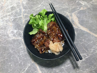 Grilled pork rice with green vegetable and Pickled ginger in the black bowl, decorate with black chopsticks on the marble table.