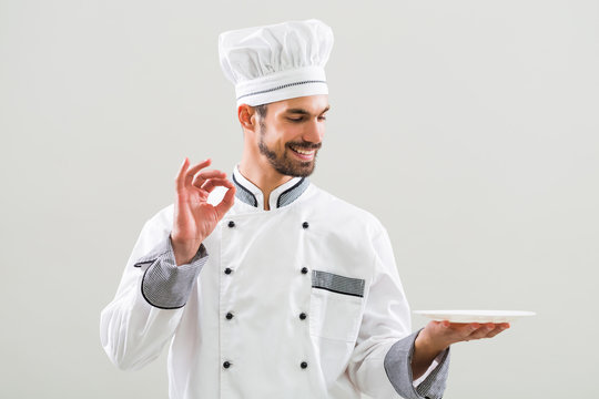 Smiling  chef is holding plate and showing ok sign on gray background.
