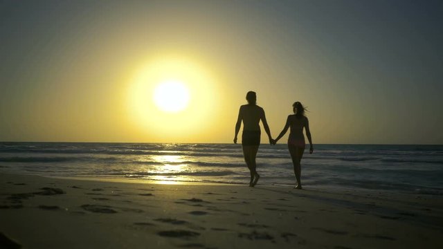 Young couple is holding hands and walking to sunset on the golden sandy beach. Two hipster in love, walks, dreams, relaxes at amazing beautiful sunset. Ocean waves washes feet of beautiful couple