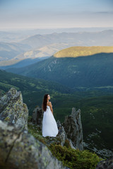 Young woman in long dress standing on the cliff edge. Beautiful young long hair woman posing in high mountain scenery. Rocky mountain